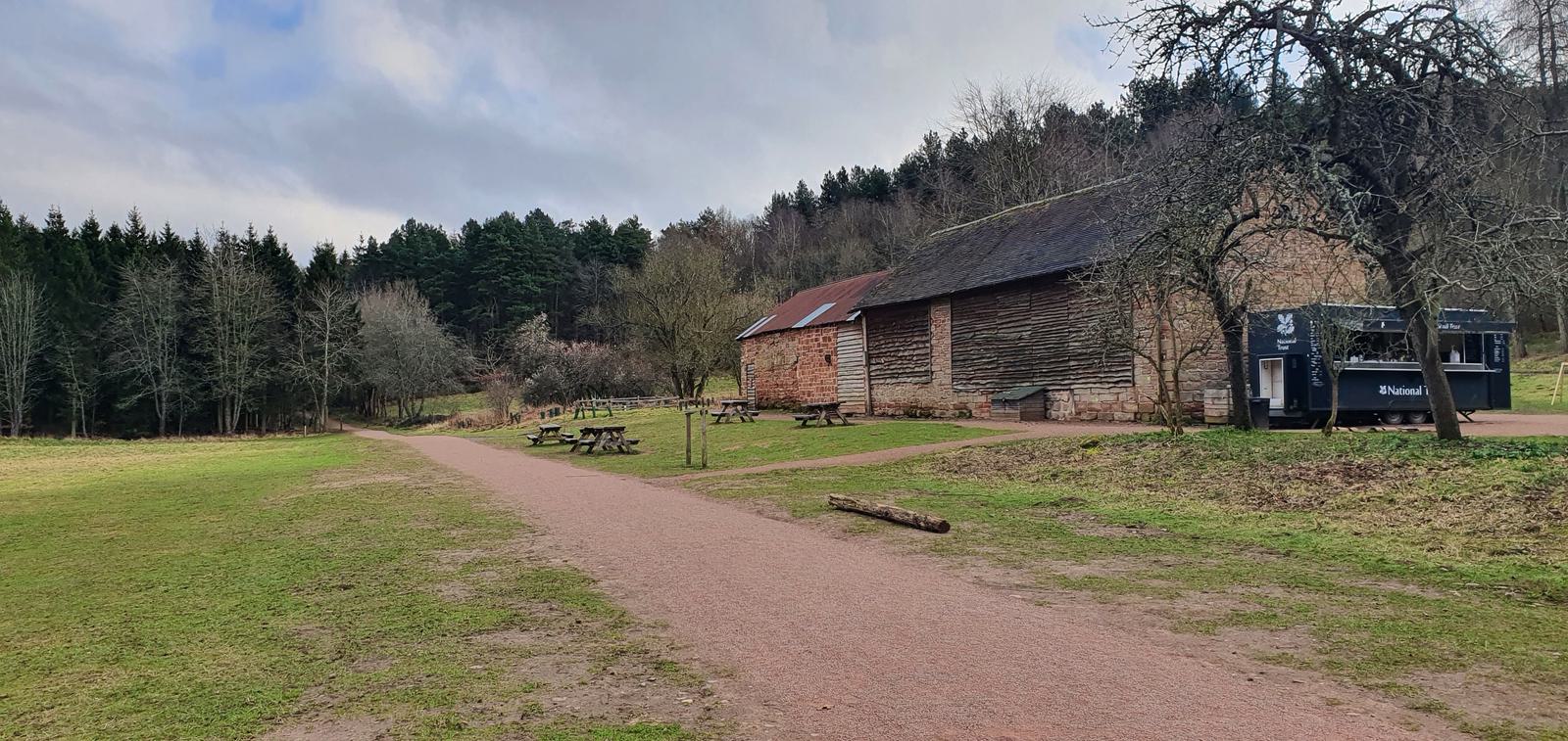 A scenic outdoor setting with a gravel path, rustic brick buildings, picnic benches, and a food truck. Trees line the background, and the sky is partly cloudy. The area appears tranquil and inviting.