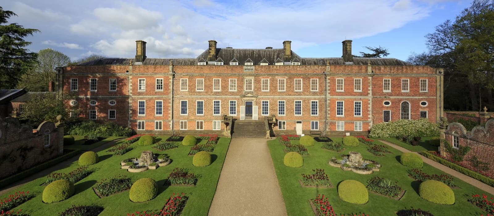 A large, symmetrical brick mansion with multiple windows and chimneys stands behind an ornate garden with trimmed hedges, pathways, and flowerbeds. The sky is partly cloudy, and tall trees are seen in the background.