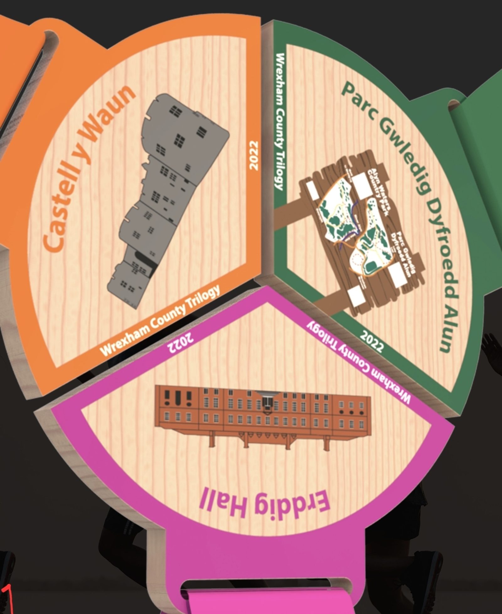 A circular wooden plaque divided into three parts, each dedicated to a landmark: Castell y Waun with an illustration of a castle, Parc Gwledig Dyfroedd Alun showing a park scene, and Eddig Hall featuring a building. Each segment is labeled "Wrexham County Trilogy" with the year 2022.