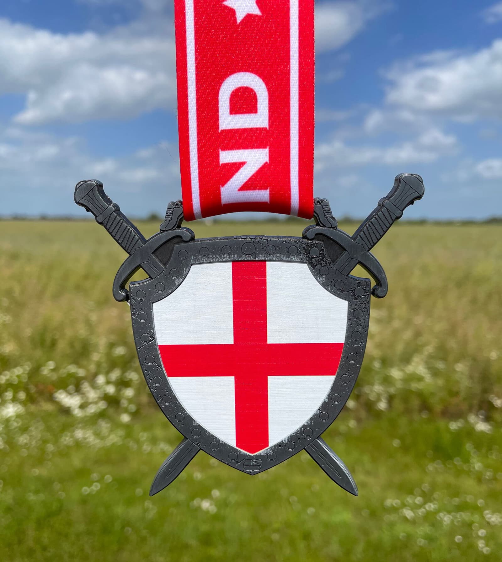 A medal featuring a shield with the red cross of St. George, representing the flag of England. The shield is flanked by two crossed swords and attached to a red ribbon with white text. In the background, there is a grassy field under a partly cloudy sky.