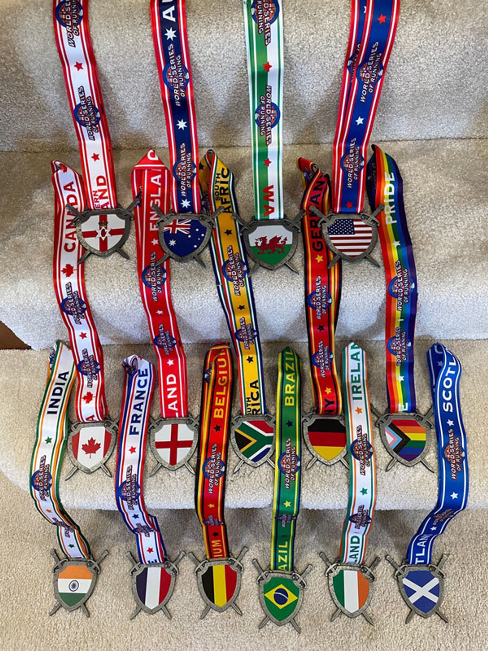A collection of medals with various national flags are displayed on beige carpeted stairs. Each medal is attached to a ribbon printed with the name and flag of a different country, including India, Canada, France, Australia, Belgium, South Africa, USA, and more.