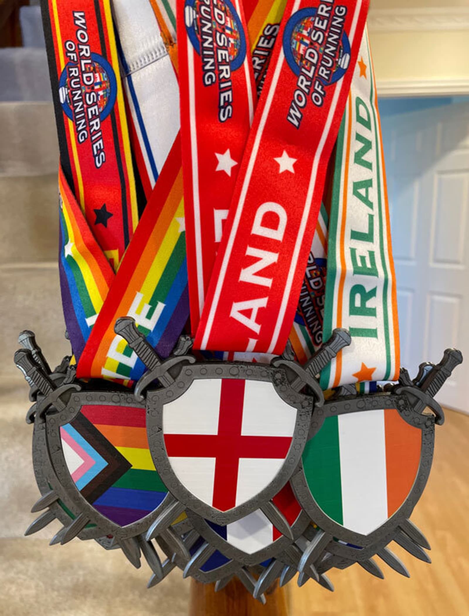 A set of medals with shields bearing various flags, including the United Kingdom's flag and Ireland's flag. The ribbons display text such as "World Series of Running." The shields feature crossed swords. The medals are displayed against a blurred indoor background.