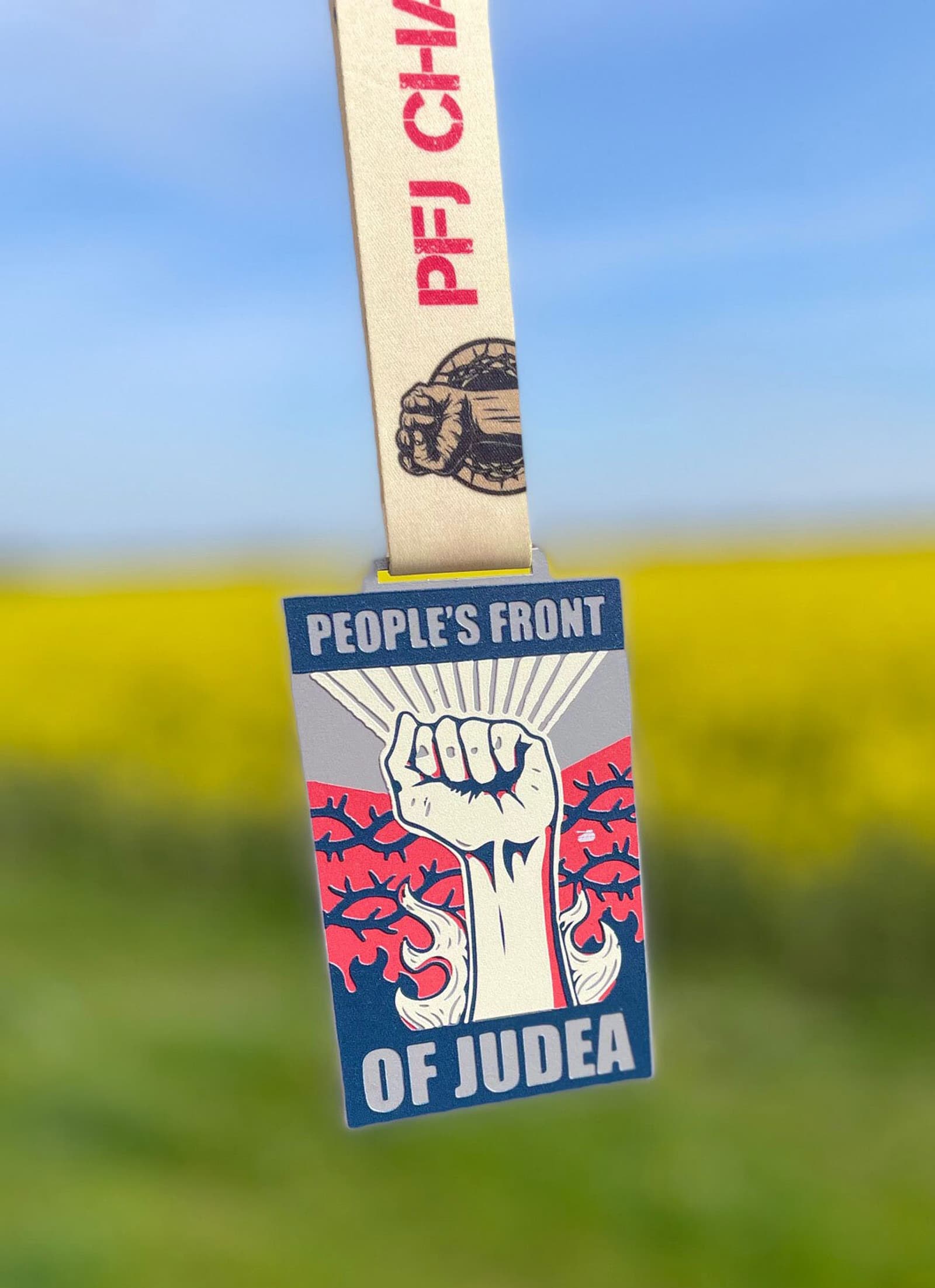 A medal with a fist and rays design, reading "People's Front of Judea," hangs from a beige ribbon inscribed with "PFJ CHAMPION" and a fist emblem. The background shows a blurred field with a blue sky, emphasizing the raised fist symbol.