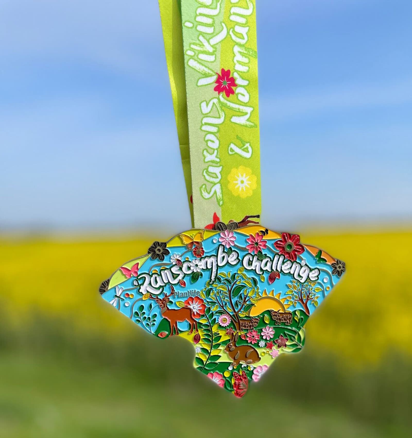 A vibrant, colorful medal with intricate floral designs and the text "Zionscombe Challenge" is attached to a green ribbon. The ribbon also has text and flower designs. The background shows an out-of-focus yellow field under a blue sky.