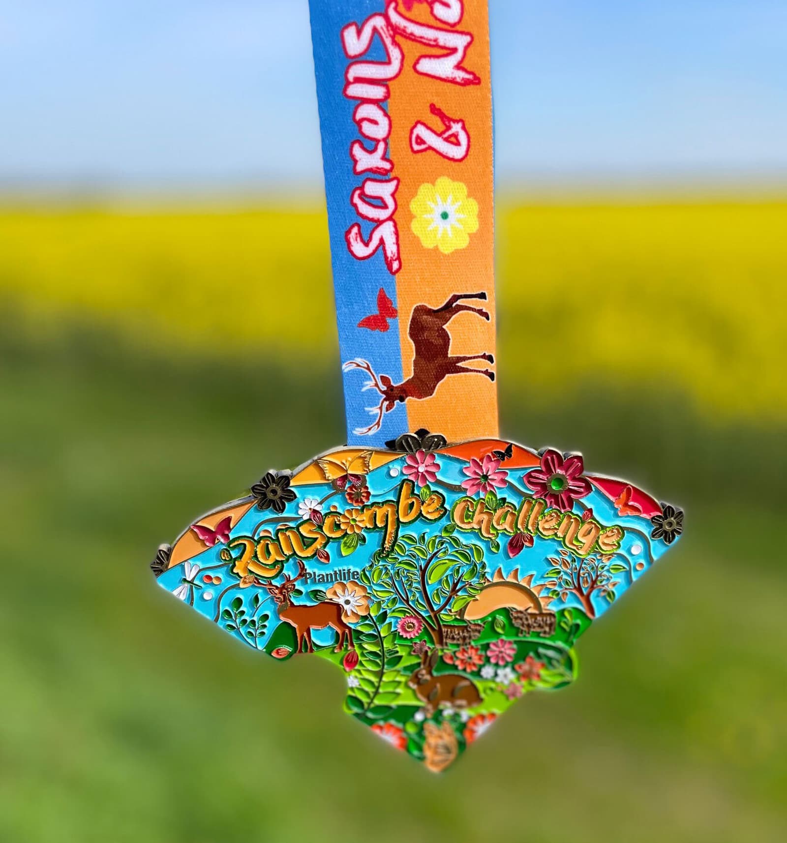 A vibrant, intricately designed medal hangs from a multicolored ribbon with "Saxons & Normans" printed on it. The medal reads "2015 Jungle Challenge" featuring jungle imagery and bright colors. The background is a blurred field of yellow flowers under a blue sky.
