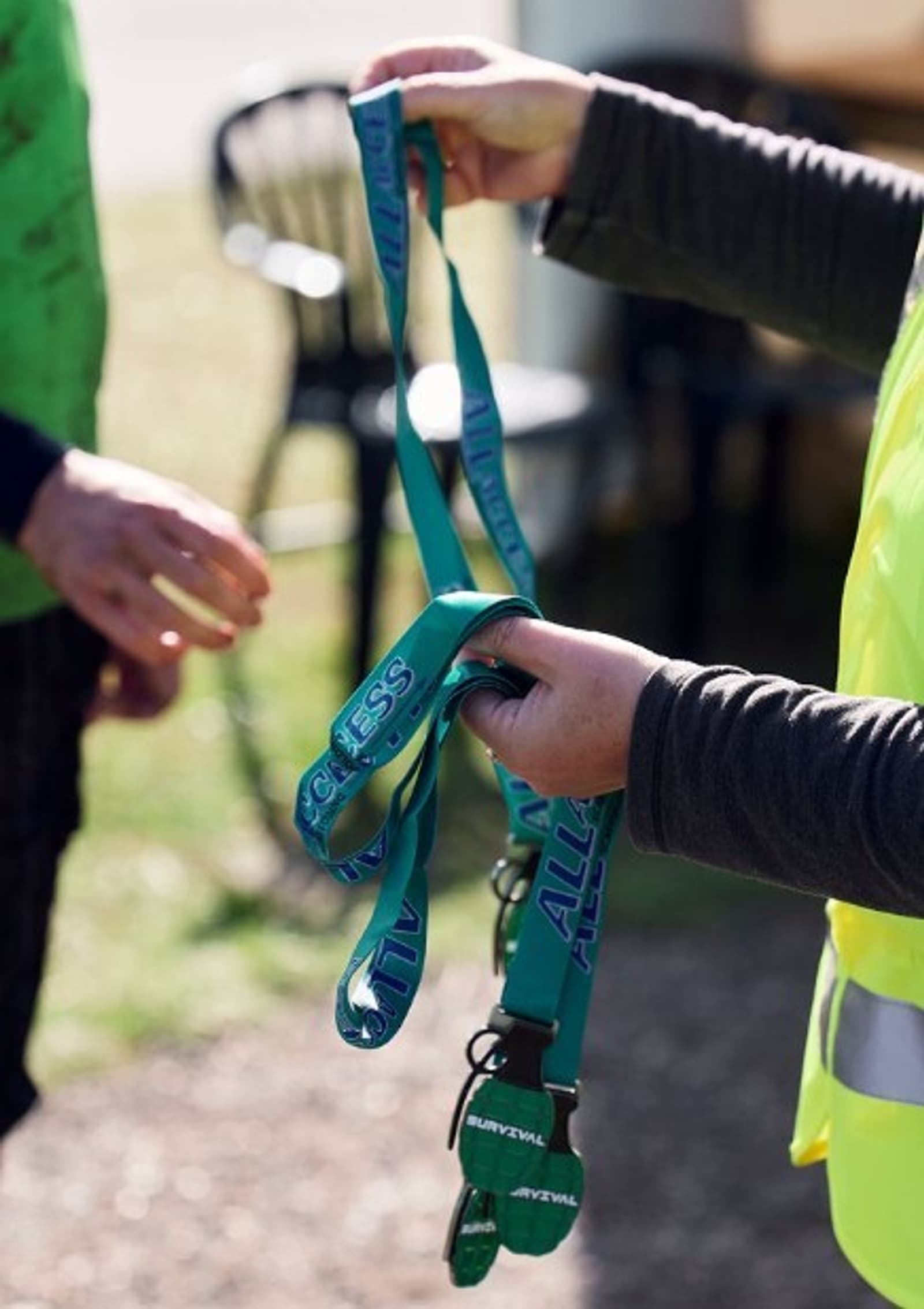 A person wearing a fluorescent yellow vest is handing blue lanyards, which say "ALL ACCESS," to someone else. In the background, a partially visible person is wearing a green top. The scene appears to be outdoors with some chairs in the distance.