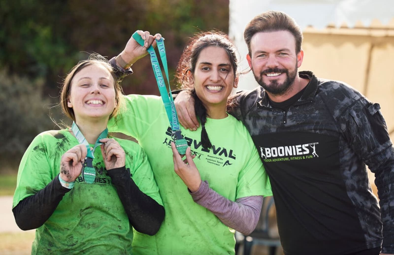 Three individuals standing together and smiling. Two women on the left, wearing green shirts, are holding medals. The man on the right, in a black shirt with a "BOONIES" logo, has his arm around the shoulder of the middle woman. They appear to have completed an outdoor event.