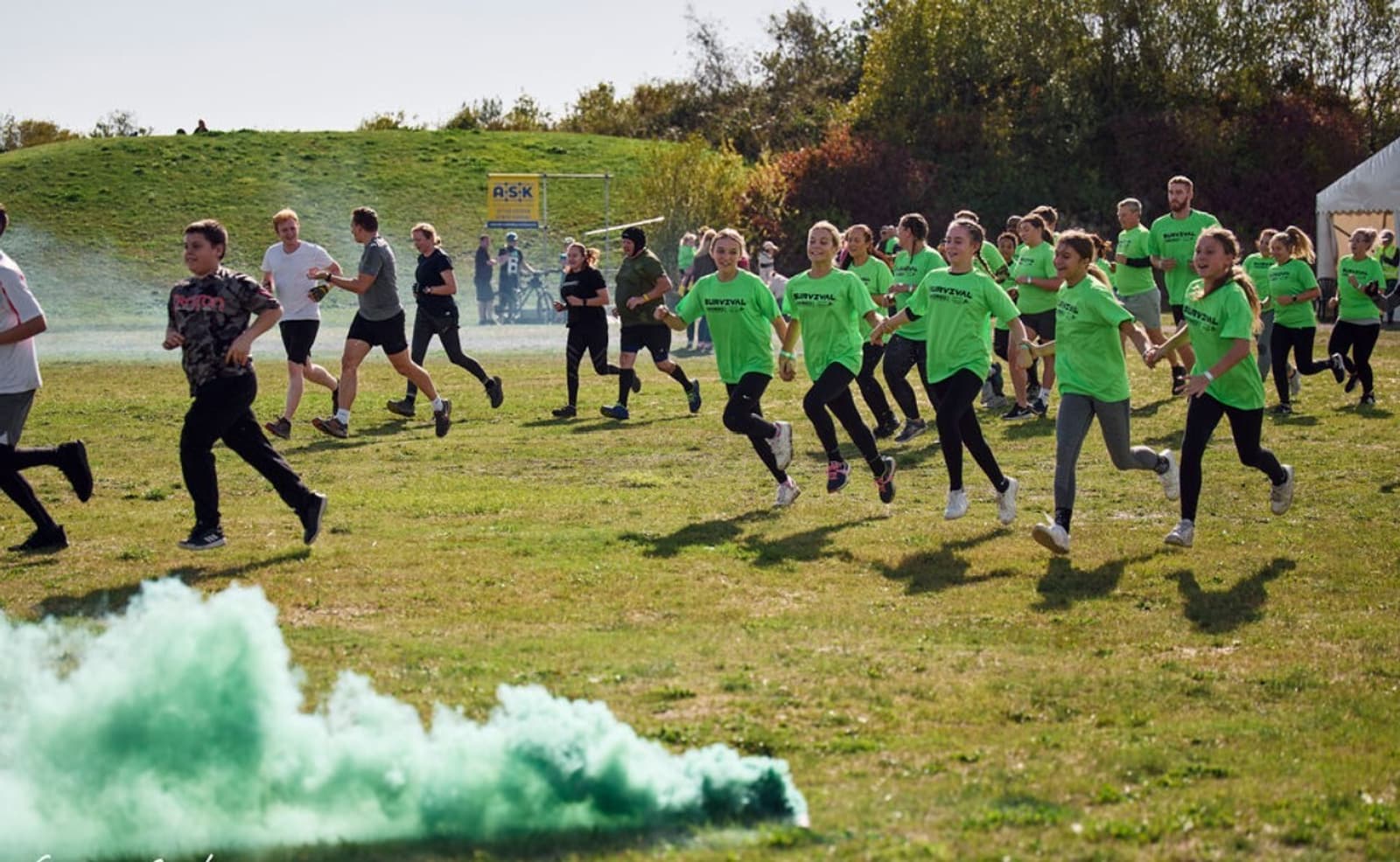 A group of people of various ages, many wearing bright green shirts, are running together in an outdoor grassy area. Green smoke is visible on the ground, and trees and a tent can be seen in the background. The atmosphere appears lively and energetic.
