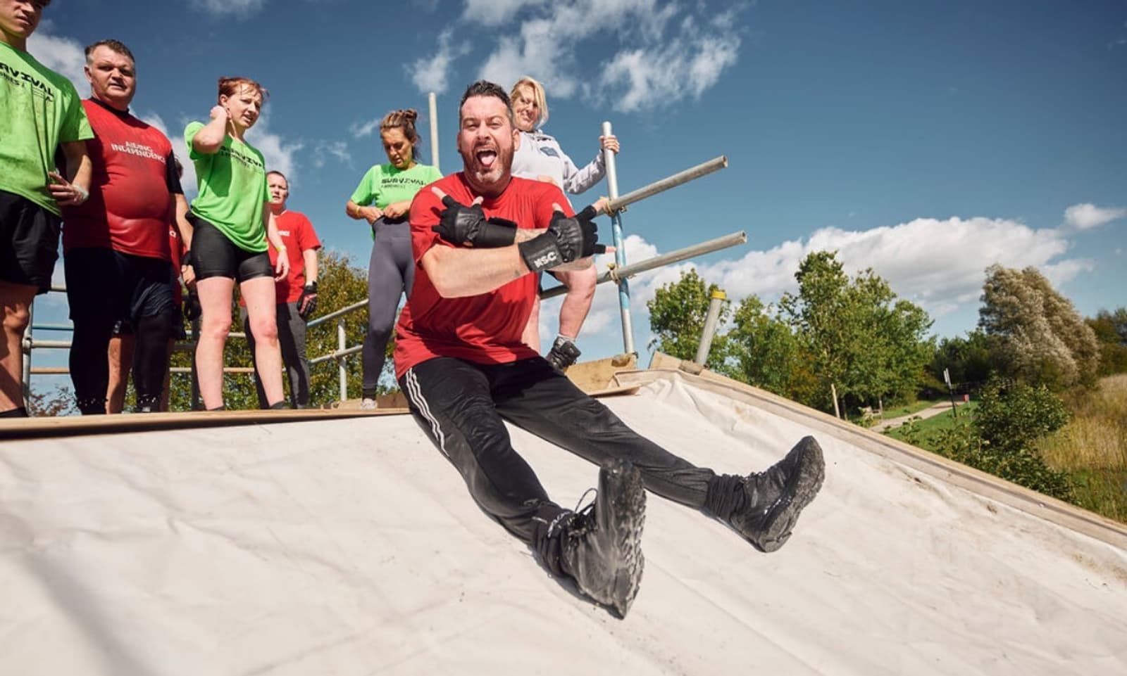 A man in a red shirt and black pants slides down a steep incline, smiling playfully with his tongue out. Behind him, a group of people in athletic wear watch and wait their turn. The scene is set outdoors under a clear blue sky with a few clouds.