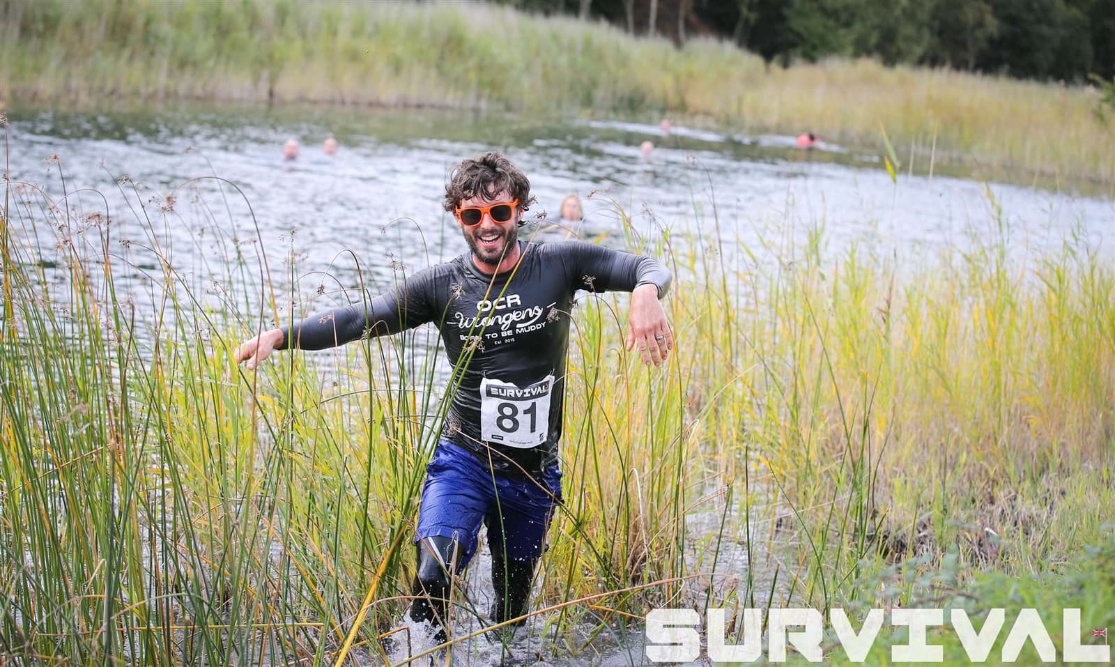 A man wearing a black shirt and orange sunglasses is wading through water and tall grass, smiling, during an outdoor survival event. He is participant number 81 and other participants can be seen in the water behind him. The word "SURVIVAL" is displayed in the foreground.