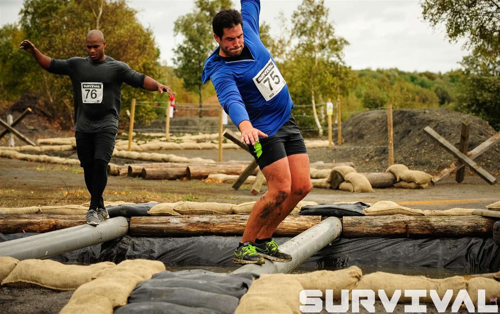 Two participants in numbered bibs navigate a balance obstacle during an outdoor survival challenge. One man in a blue shirt and black shorts balances on beams while another in gray follows behind. Sandbags and log barriers are visible, with overcast skies overhead. "SURVIVAL" is written at the bottom right.
