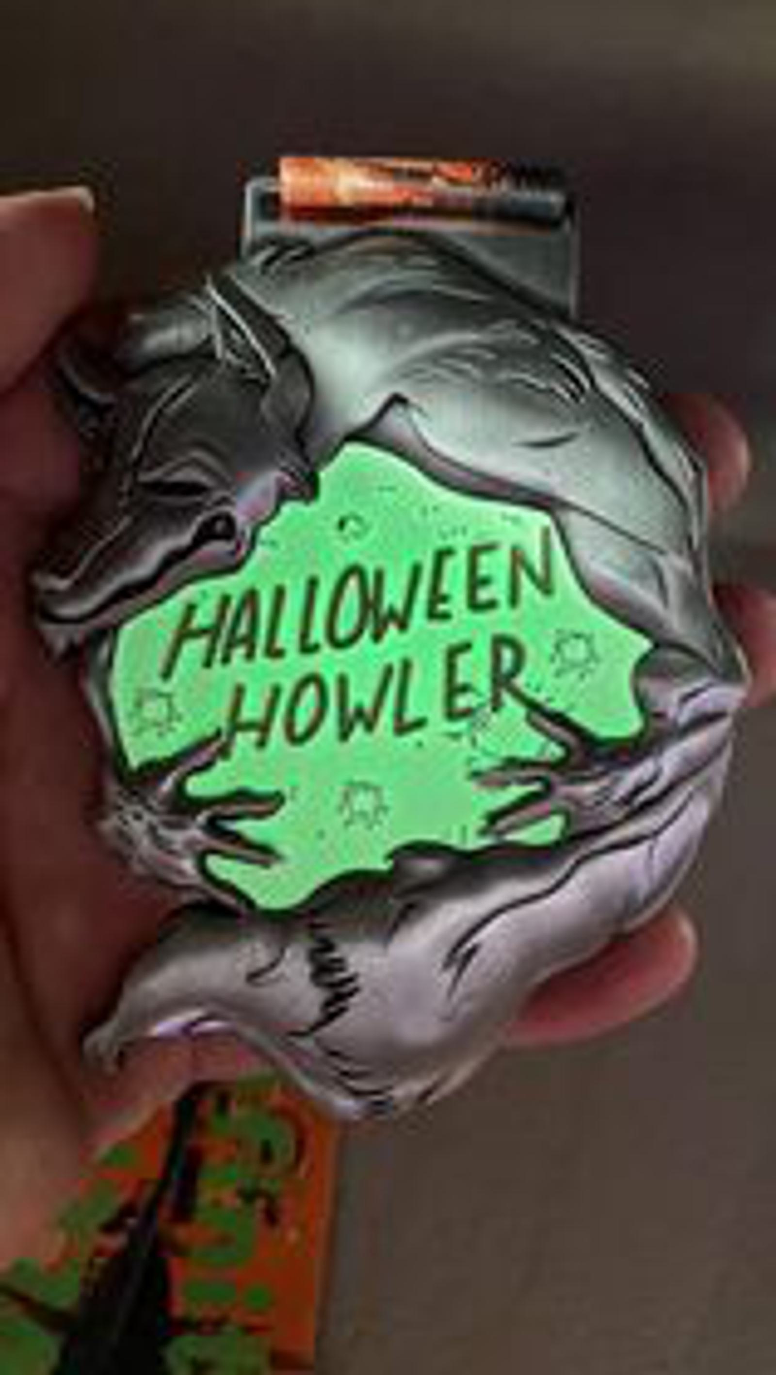 A hand holds a spooky-themed medal with a glowing green center that reads "Halloween Howler." The medal features creepy hands and a wolf howling at the top, enhancing the Halloween motif.