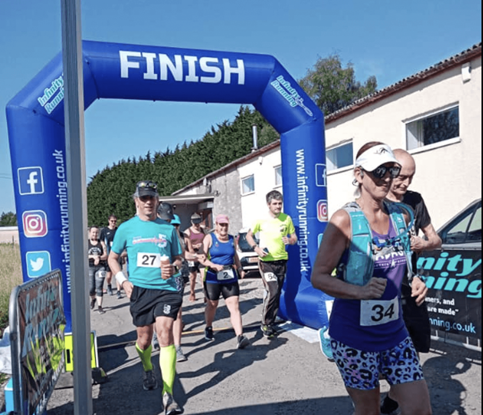 Runners, wearing athletic gear and numbered bibs, cross a blue "FINISH" arch at an outdoor race event on a sunny day. A man in a teal shirt and a woman in a purple top are prominent. Other participants and spectators are visible in the background.
