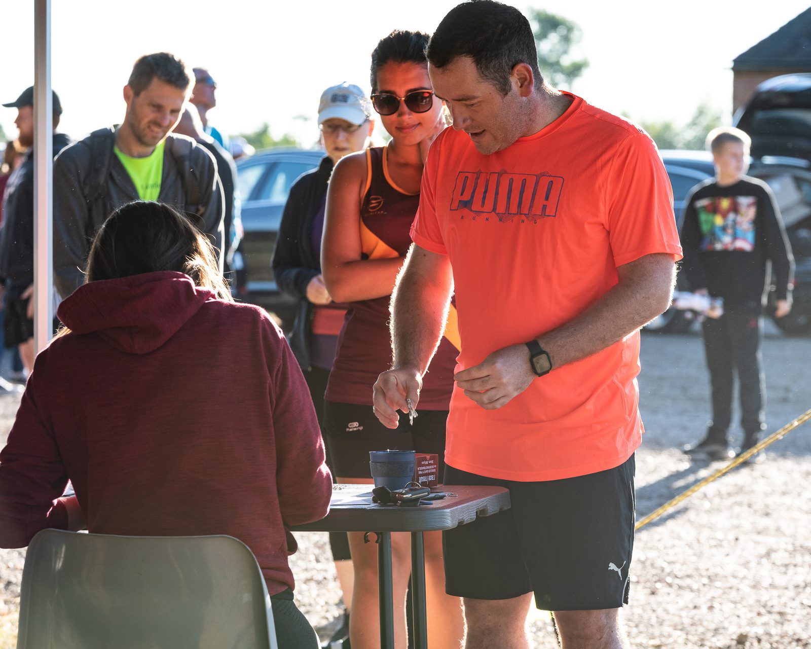 A man in an orange shirt signs paperwork at an outdoor registration table, while a woman in a maroon shirt assists him. Another woman in sunglasses and a maroon tank top stands in line behind him. Several other people are visible in the background.