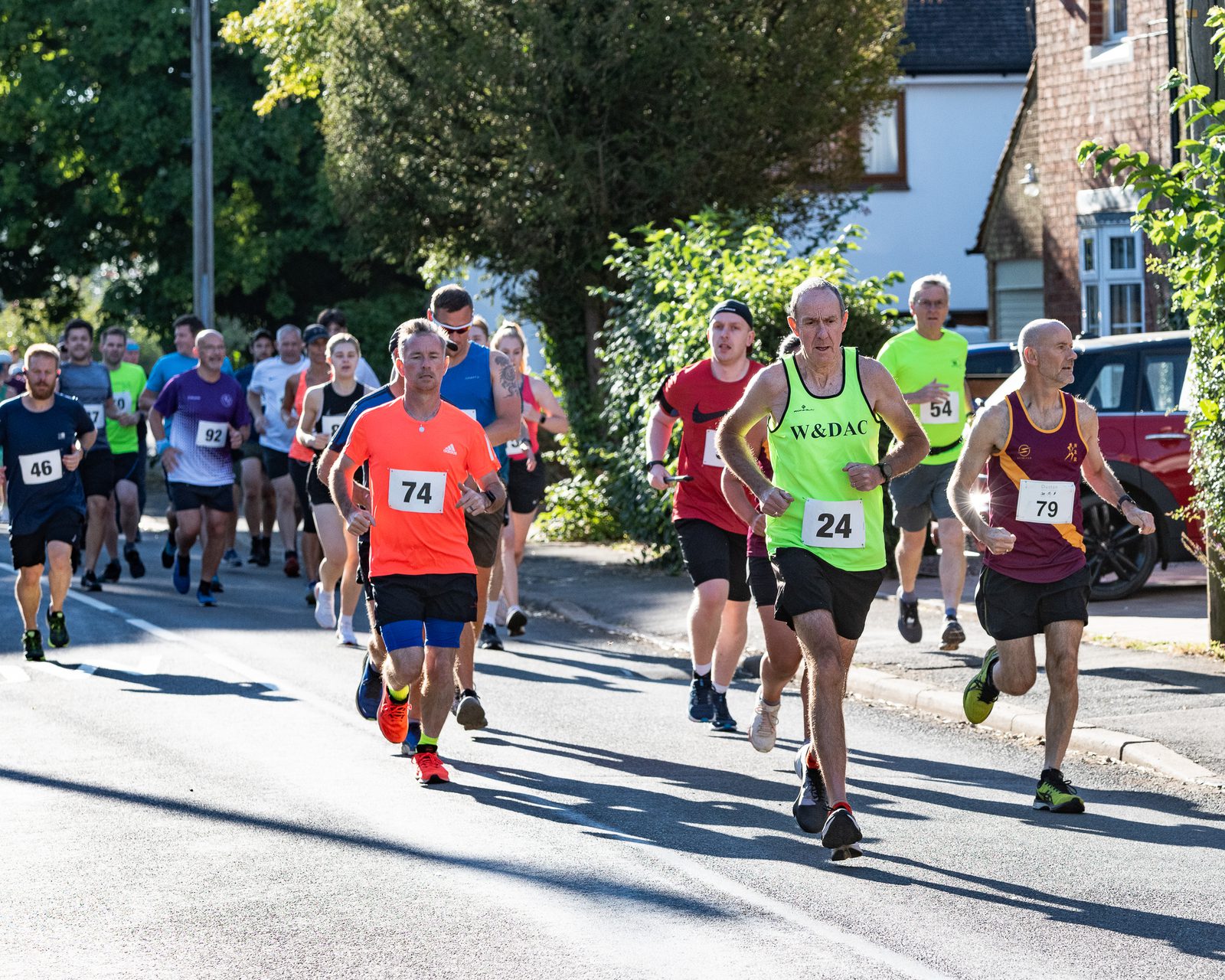 A group of runners participate in a road race in a residential area. The runners, wearing numbered bibs and various brightly colored athletic gear, move forward under the sunlight. Trees and houses line the street, with more runners visible in the background.