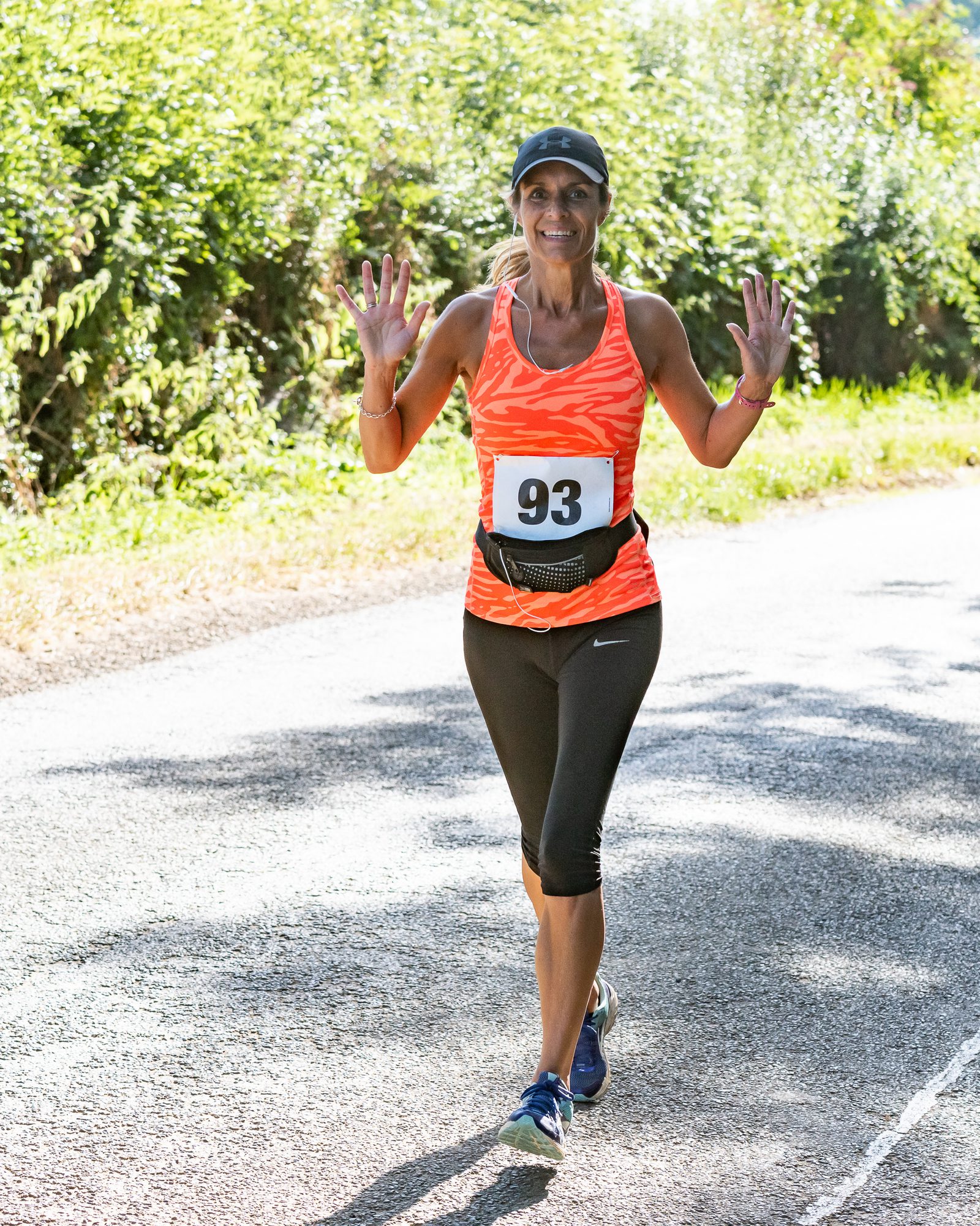 A woman wearing a bright orange tank top, black capri leggings, and a black cap with bib number 93 is running on a shaded road surrounded by greenery. She is smiling and has both hands up, seemingly waving or showing ten fingers.