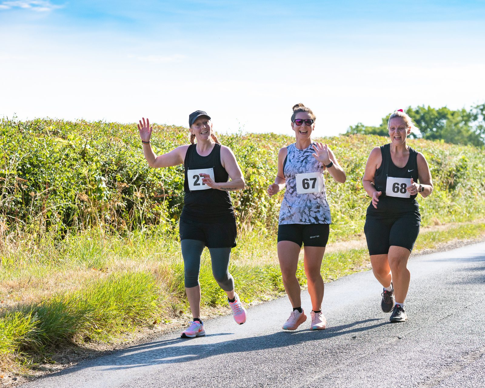 Three women participants with bib numbers 27, 67, and 68 are running and smiling in an outdoor race on a sunny day. They are wearing athletic gear and passing by green bushes along the roadside. The woman on the left is waving while running.