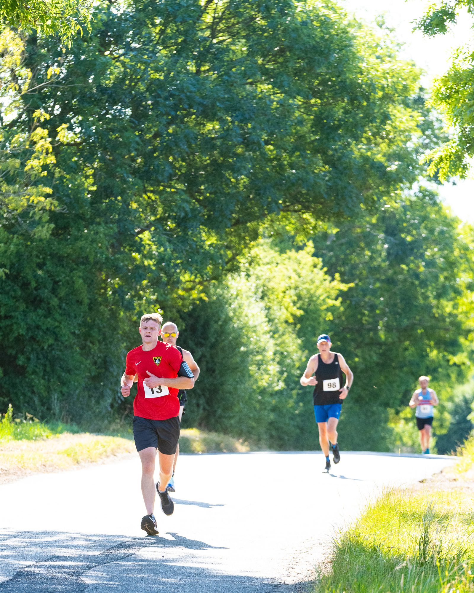 A group of runners race on a sunlit road surrounded by lush greenery. The leading runner wears a red shirt and is followed closely by others in athletic attire. The bright, clear weather casts shadows and highlights the vibrant outdoor setting.