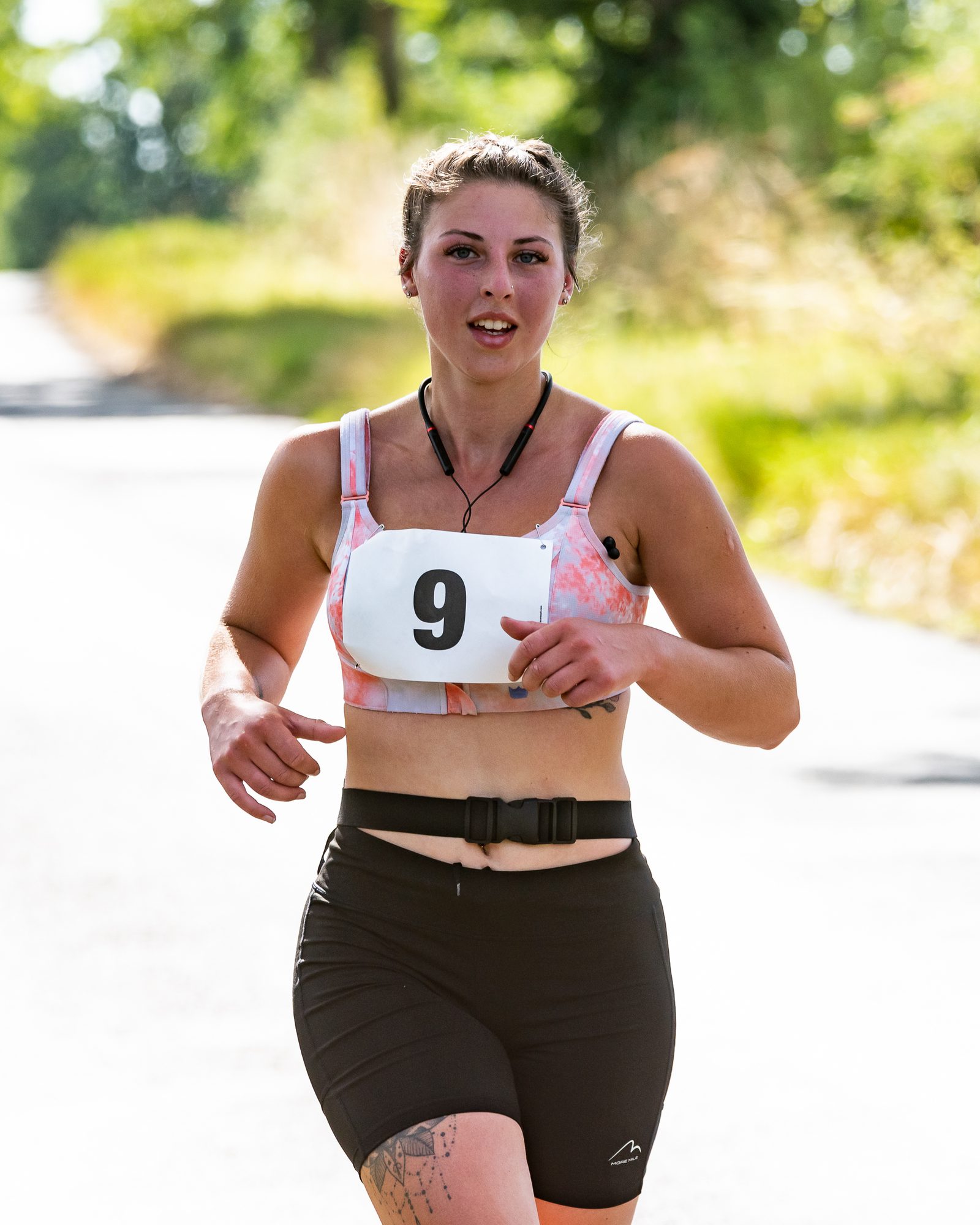 A woman participating in a road race wears a sports bra and shorts. She has a race bib with the number 9 pinned to her top and is running on a sunlit, tree-lined path.