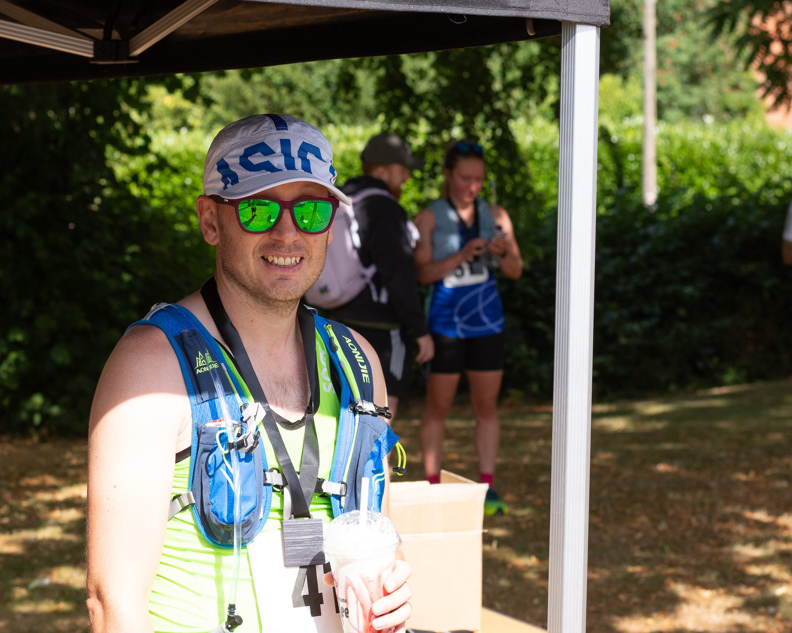 A man wearing a white and blue cap, green sunglasses, and a hydration pack smiles while holding a drink. He stands under a canopy outdoors. In the background, another person in blue activewear is looking down, and lush greenery surrounds them.