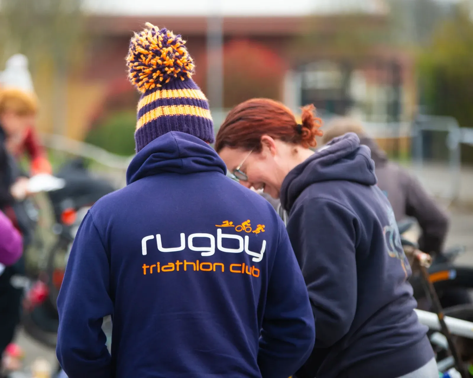 Two people facing away from the camera, one wearing a purple hoodie with "rugby triathlon club" text and a colorful knit hat with a pompom. The other person has a ponytail and eyeglasses. They appear to be at an outdoor event or gathering. Blurred background.