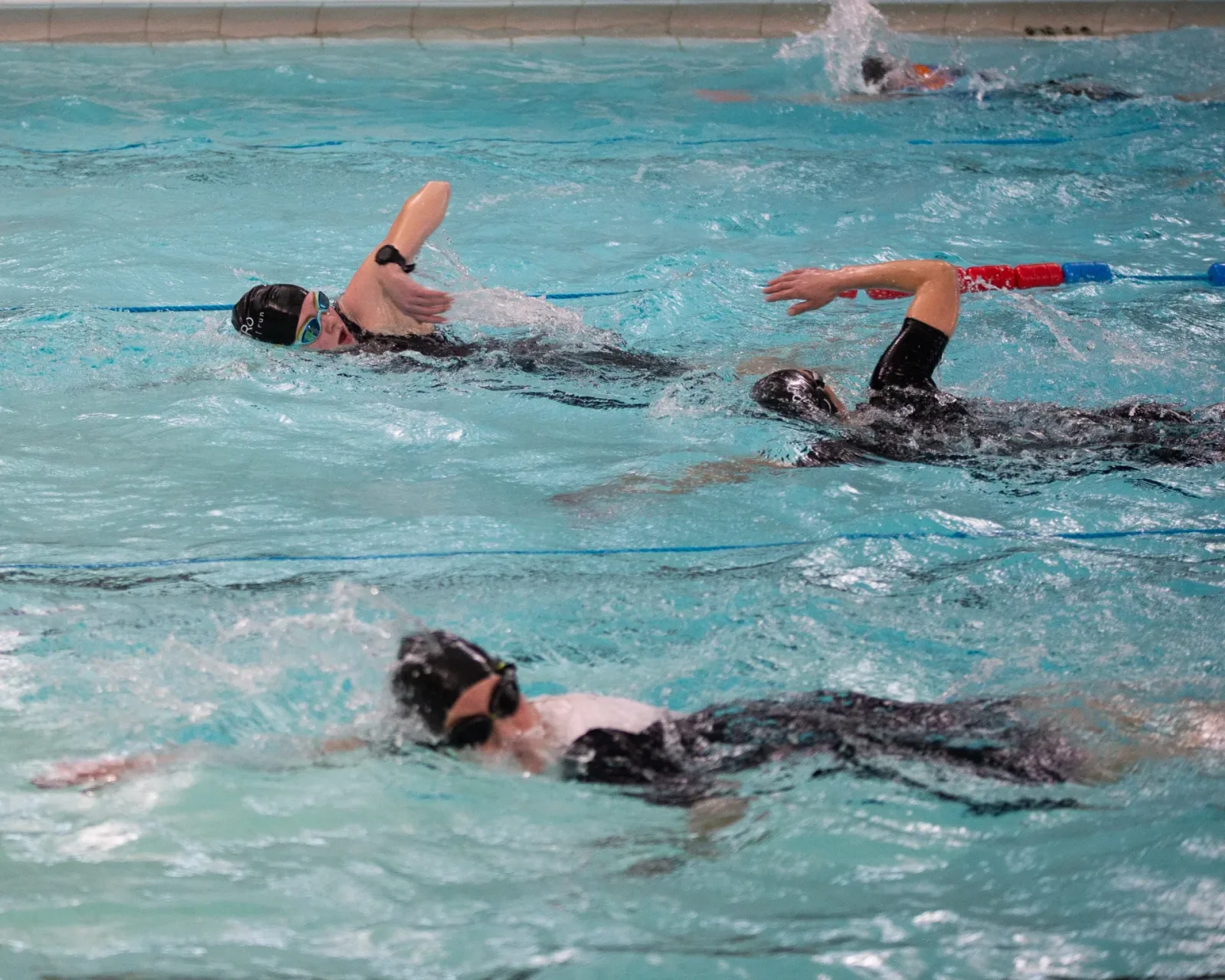 Three swimmers wearing swim caps and goggles swim freestyle in a pool. The swimmer in the foreground is in a white cap, while the others are in black caps. They appear to be in a race or training session, with clear lanes defined by floating lane markers.