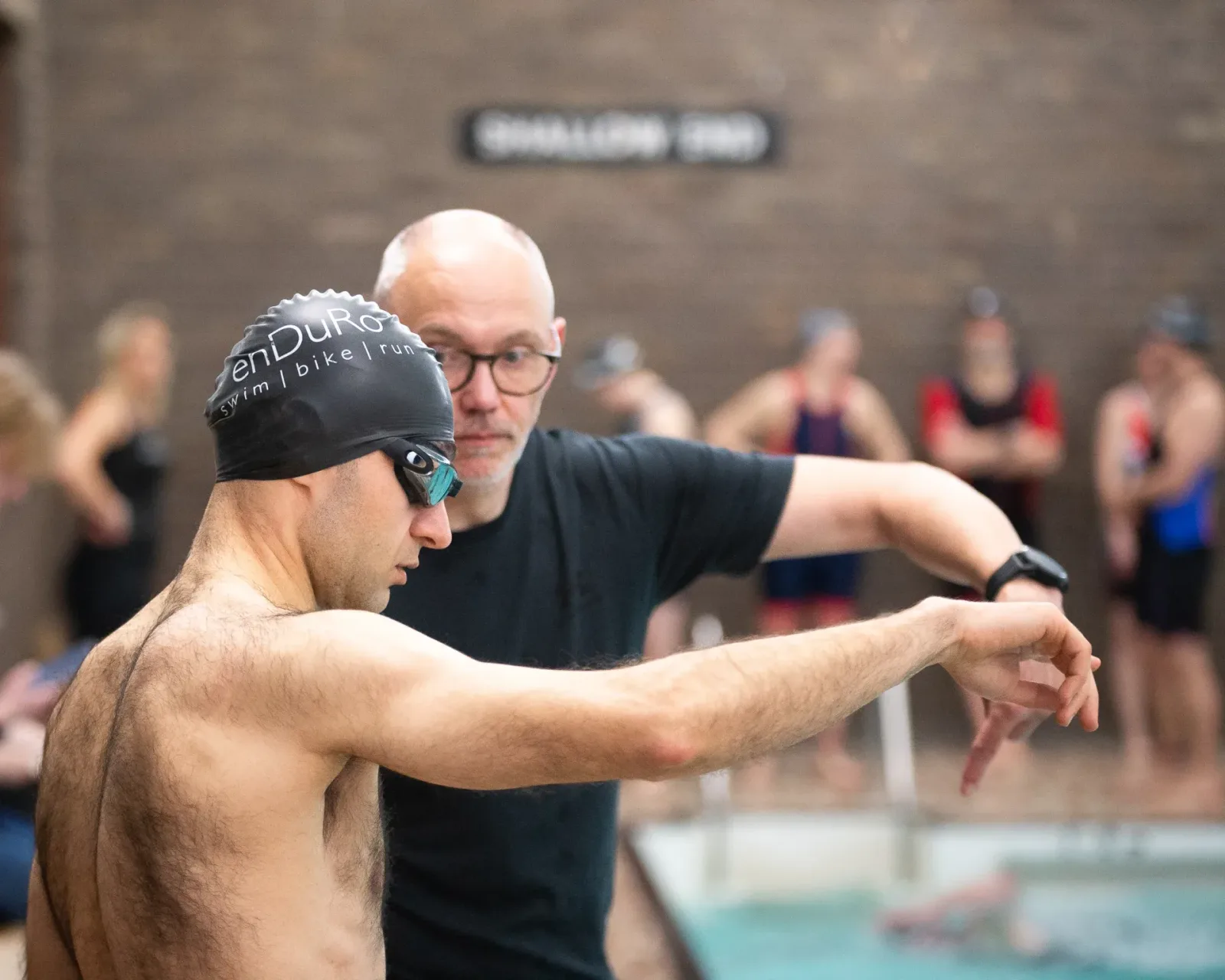 A man wearing swim goggles and a swim cap is being coached by another man in glasses by the edge of an indoor pool. Other swimmers are in the background. The coach is explaining a technique, gesturing with his hands. A "Shallow End" sign is visible on the wall.
