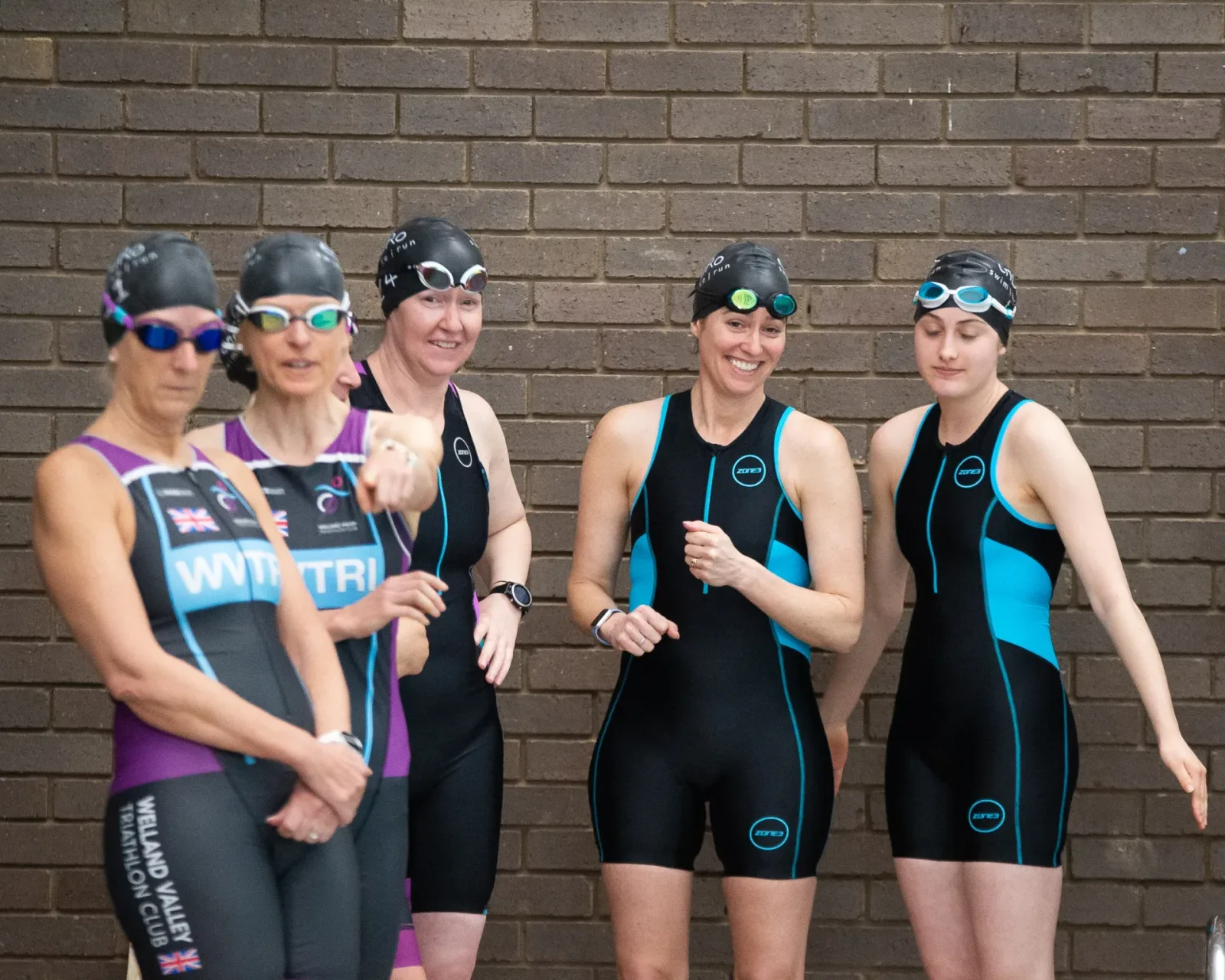 Five women in swimwear and swim caps stand in front of a brick wall. Two women on the left wear purple and black swimsuits, while the three on the right wear black and blue swimsuits. They appear to be chatting and laughing, likely before a swim event.