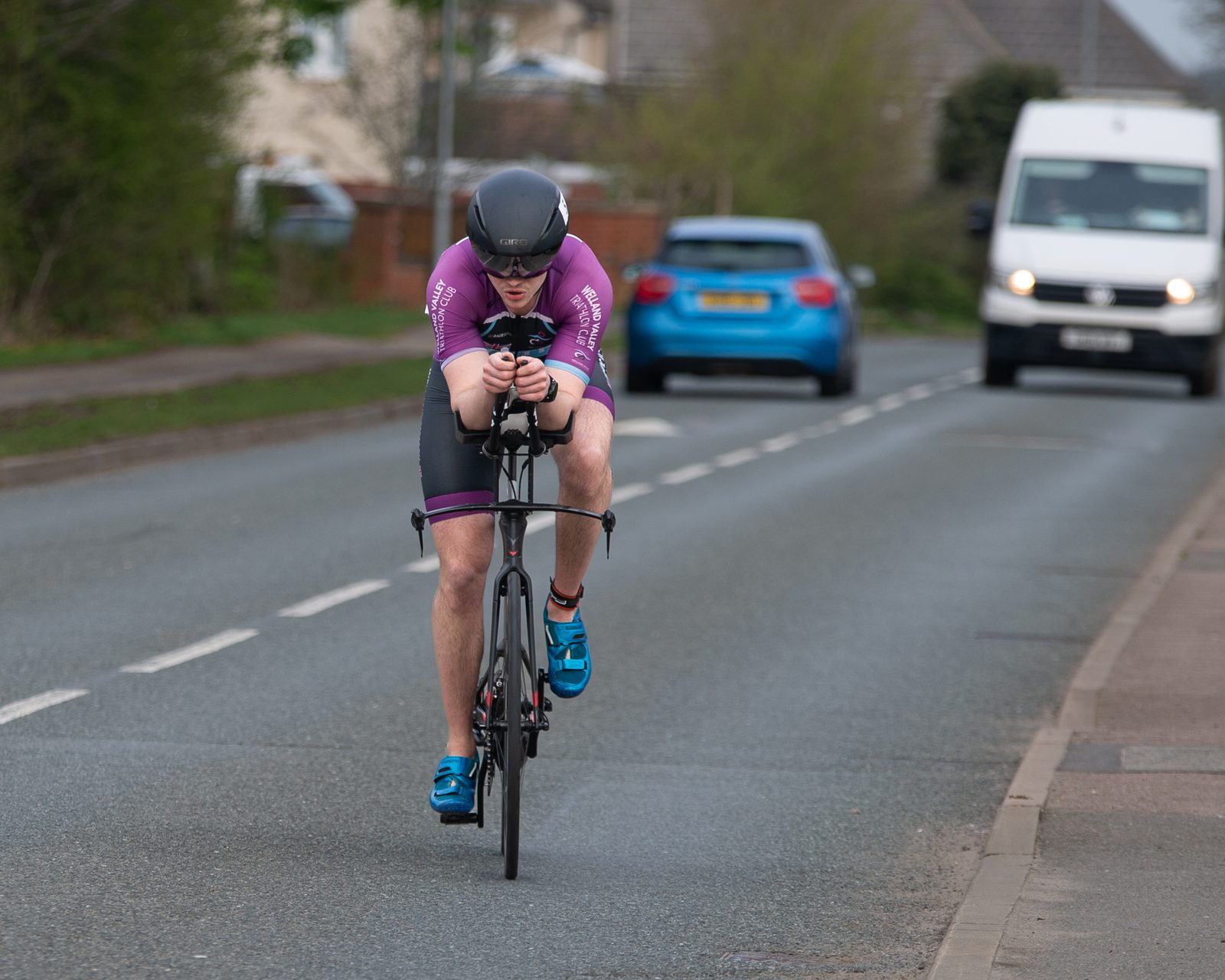 A cyclist in a purple jersey and black shorts rides a black bike on a road, leaning forward in an aerodynamic position. They wear a black helmet and blue shoes. Two vehicles are visible in the background, a blue car and a white van, on the same road.