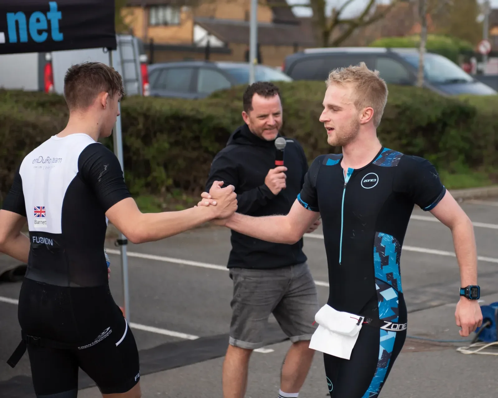 Two male athletes wearing triathlon suits and racing bibs exchange a handshake at the end of a race. One athlete wears a black and white suit with a British flag, while the other wears a black and blue suit. A man holding a microphone stands behind them, smiling.