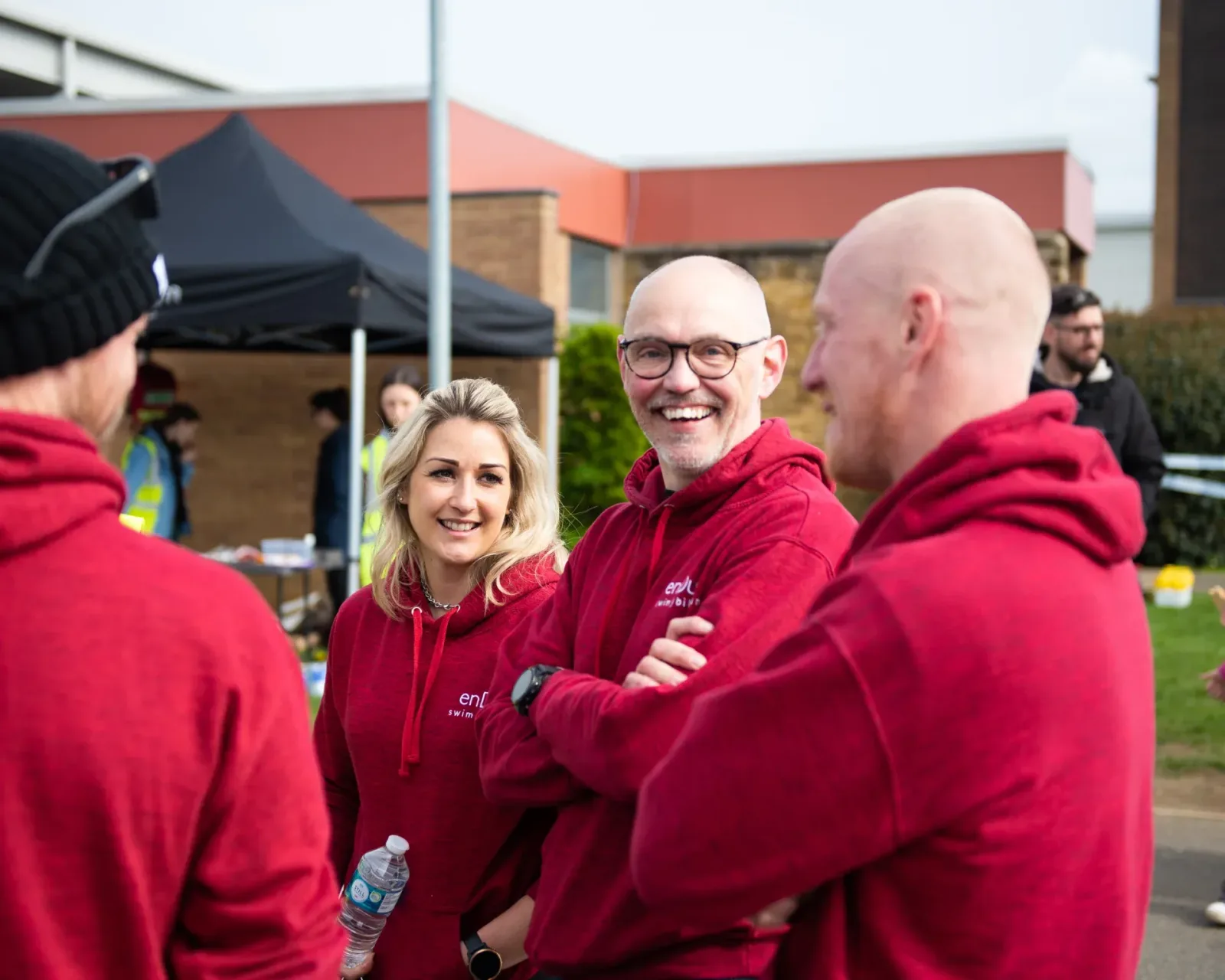 Four people standing and talking outside, three of them wearing red hoodies and one a black winter hat. They appear to be at an event with a tent visible in the background. One person is holding a water bottle, and the group looks cheerful and engaged in conversation.