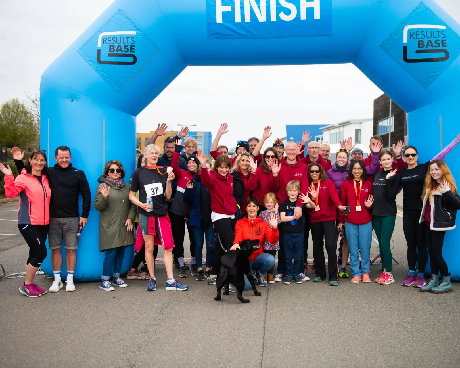 A group of people, adults and children, posing and waving under a blue inflatable finish line arch labeled "Results Base." One person in front is holding a black dog, and others around are smiling, wearing casual athletic clothing. The group looks celebratory.