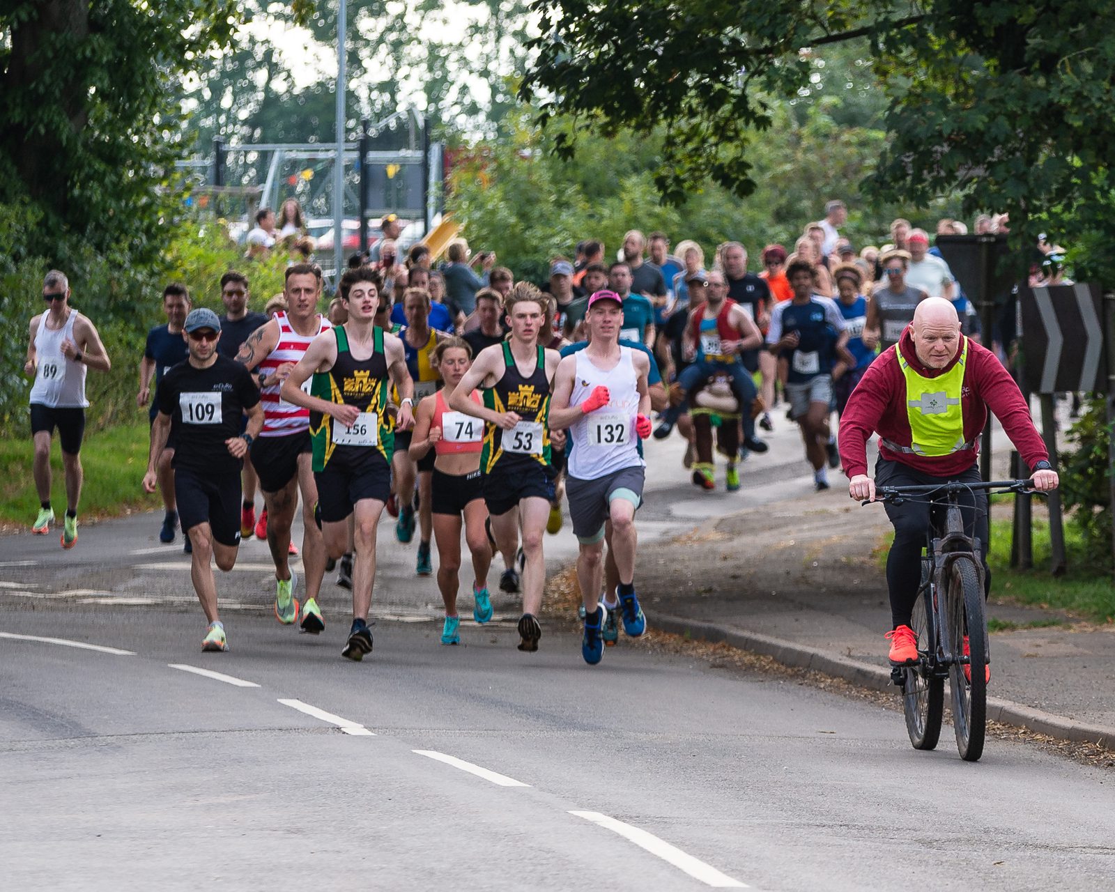 A large group of runners, many wearing numbered bibs, participate in a race on a paved road bordered by trees. A man in a red and yellow shirt rides a bicycle at the front, leading the runners. It appears to be a sunny day with clear, vibrant surroundings.