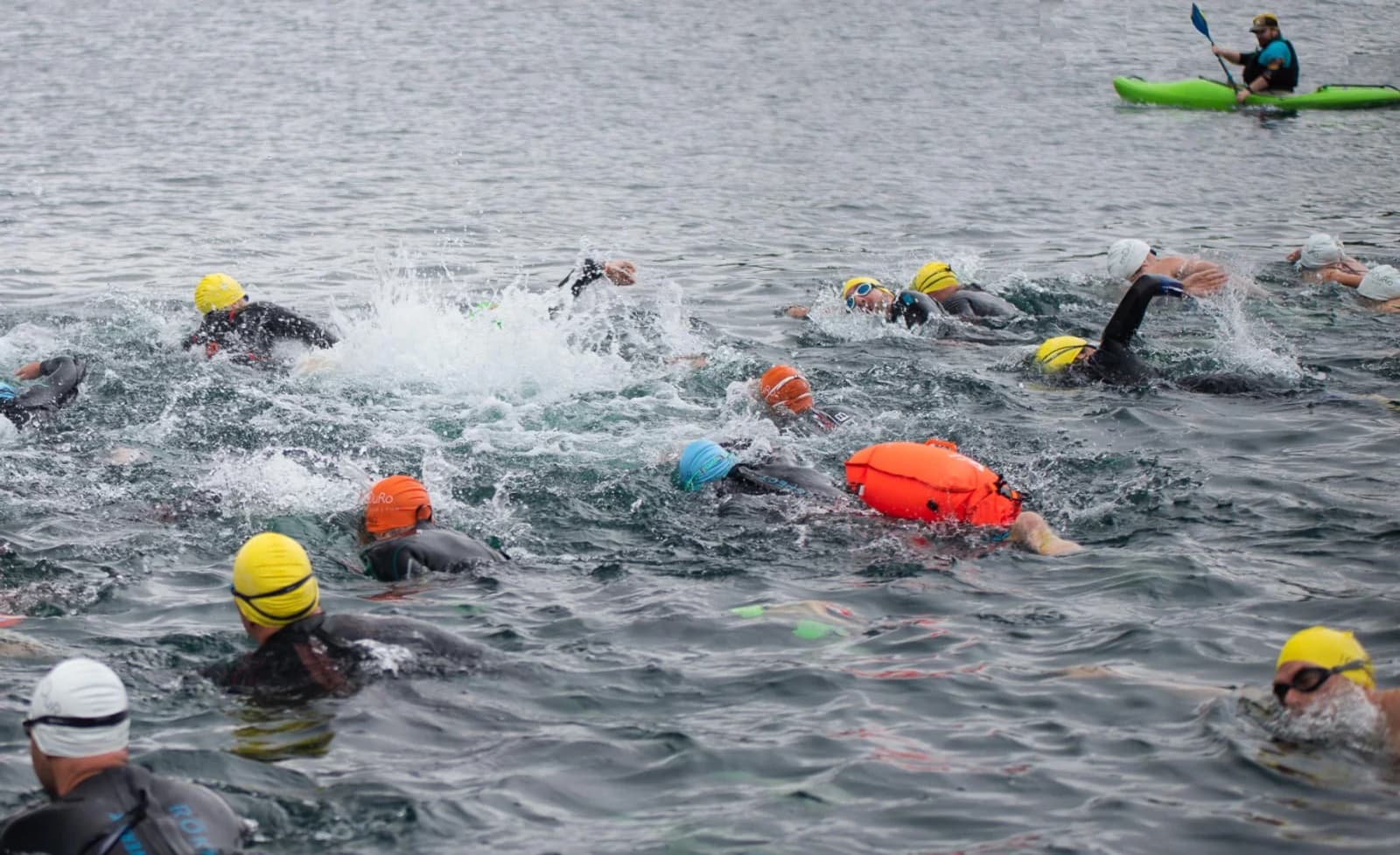 A group of swimmers wearing wetsuits and colorful swim caps participate in an open water race as a person in a green kayak observes from a distance.