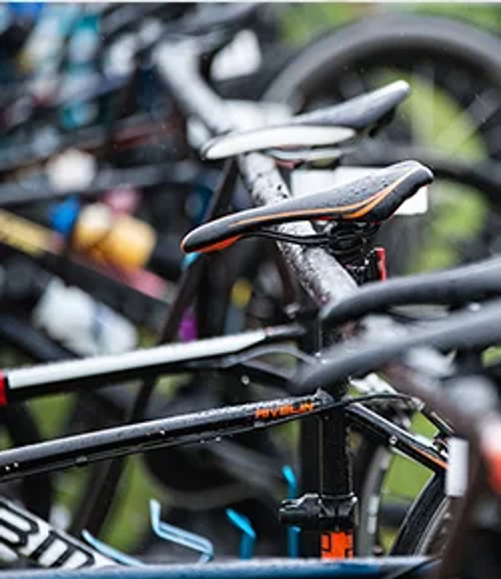 Close-up of several bicycles parked in a row, with rain droplets visible on the frames and seats, suggesting they are outdoors on a wet day. The focus is on the saddle and top tube of one bike in the foreground.