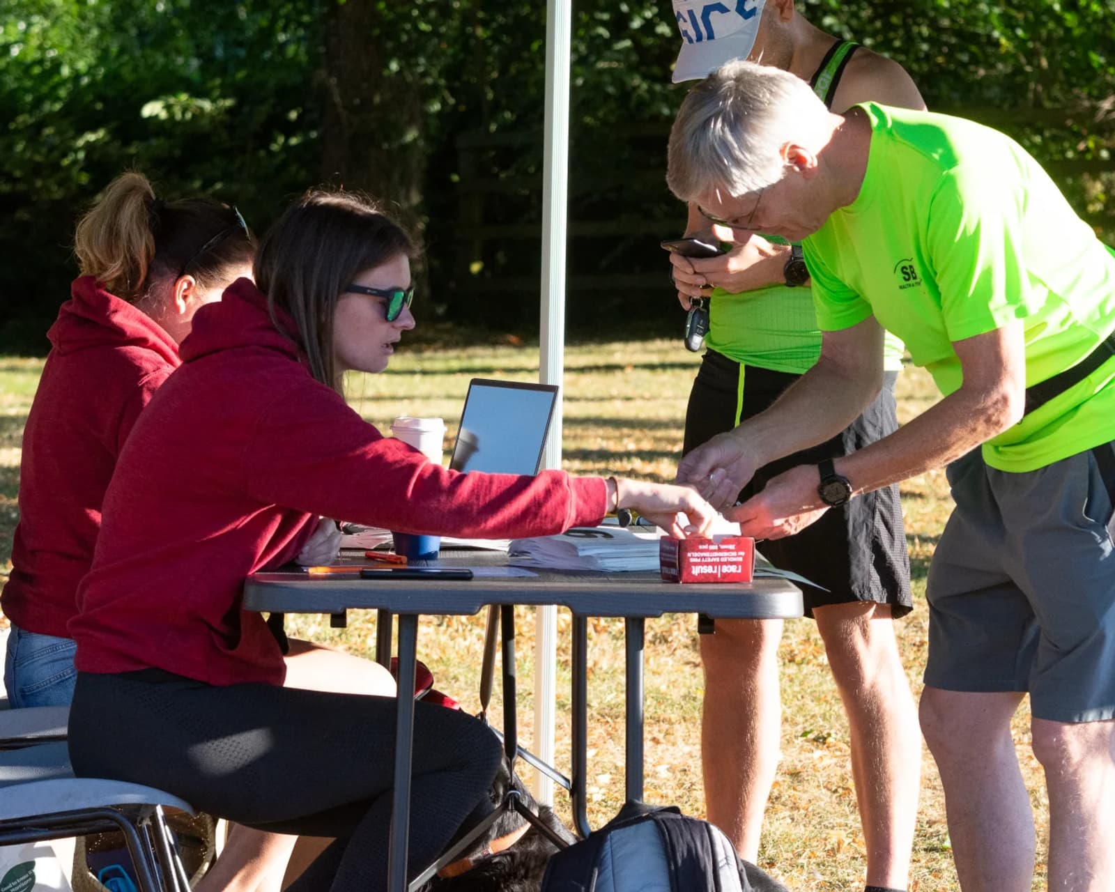 Two women sitting at an outdoor table help two men standing in front of them; one woman hands a small item to an older man in a neon green shirt while the others look on, with trees in the background.