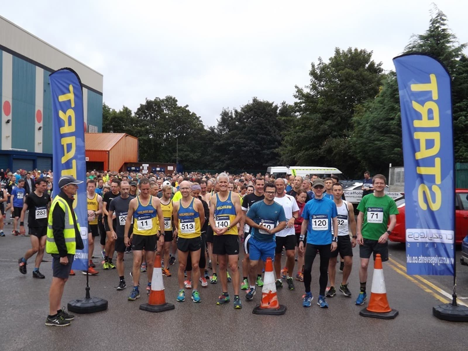 A large group of runners, wearing numbered bibs, stand at the start line of a road race. Two blue banners with the word "START" mark the beginning of the course. A race official in a yellow vest stands at the side, and trees and buildings are visible in the background.