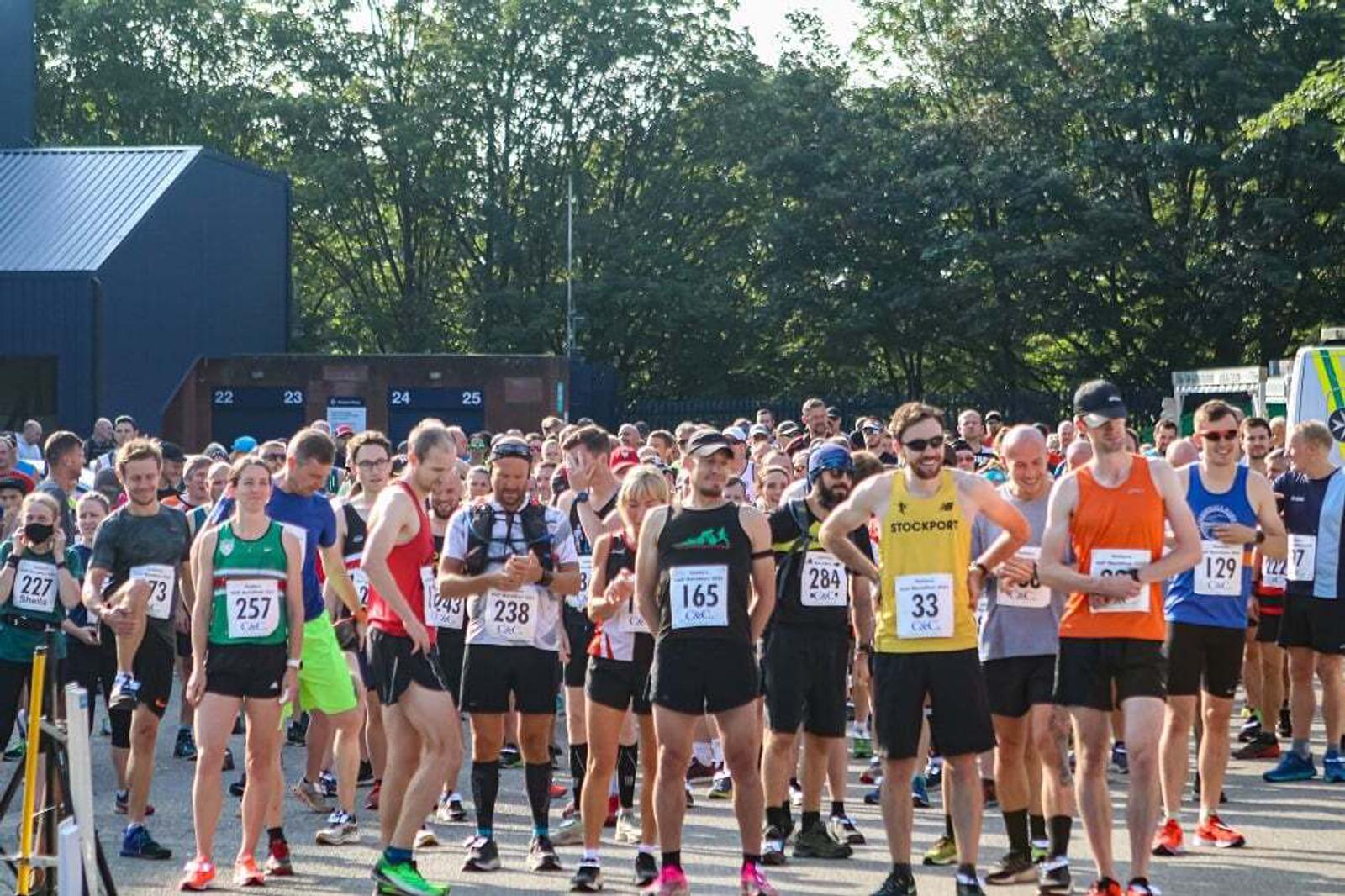 A large group of runners stands at the starting line of a race event. They are wearing athletic gear and numbered bibs. Some people are chatting while others are adjusting their gear. The backdrop includes trees, a blue building, and race facilities.