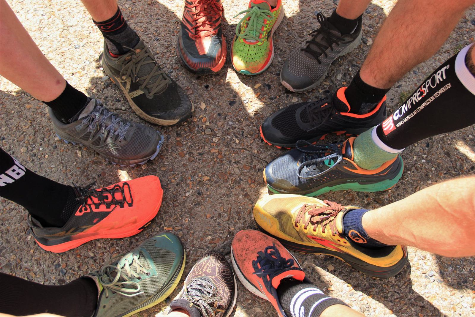 A group of people stand in a circle, showcasing their various running shoes on a sunlit gravel surface. The footwear displays a range of colors and styles, including bright orange, green, gray, and black. Some are wearing socks with branded logos.
