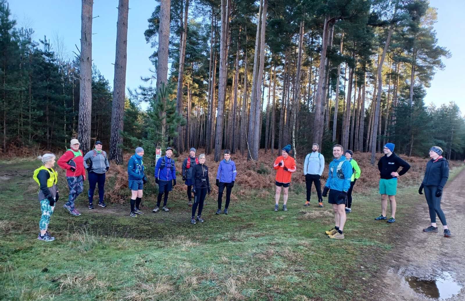 A group of people dressed in athletic and winter gear stand in a semi-circle at the edge of a forest. Tall trees and patches of grass surround them. Some individuals are talking, while others are looking around or adjusting their gear.