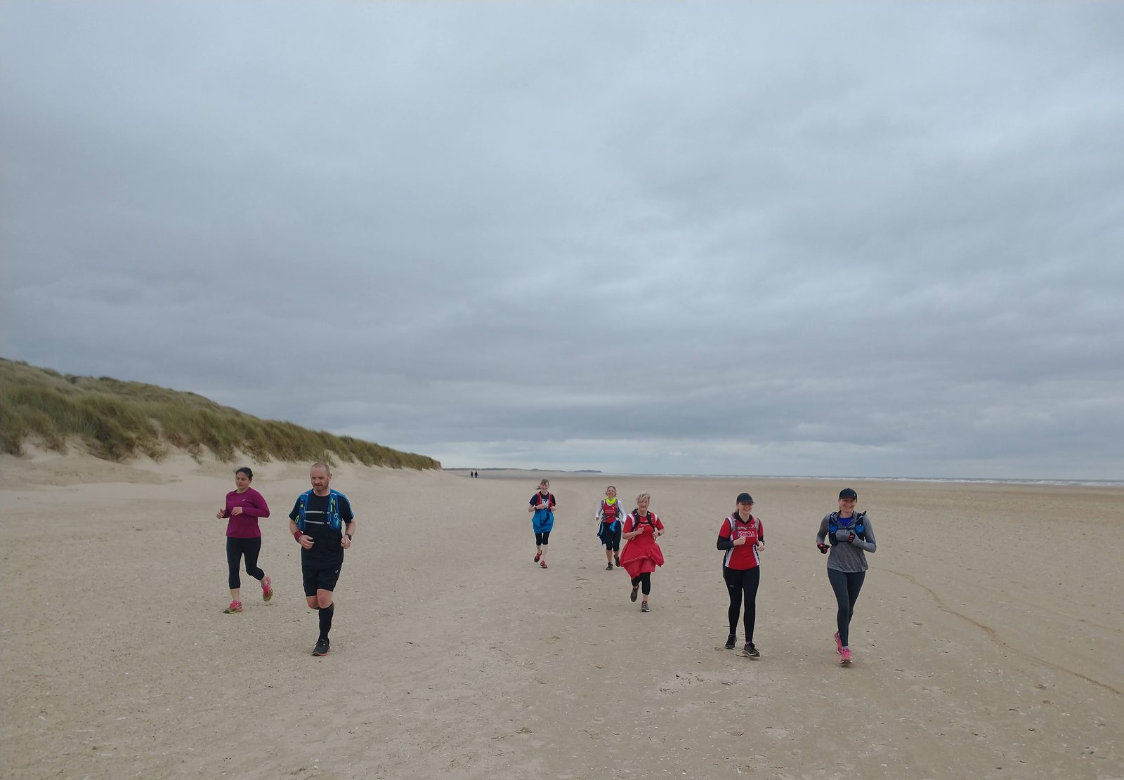 A group of eight runners in colorful athletic clothing jogs along a wide, sandy beach under a cloudy sky. Some grass-covered dunes are visible to the left, and the vast expanse of sand stretches to the right.