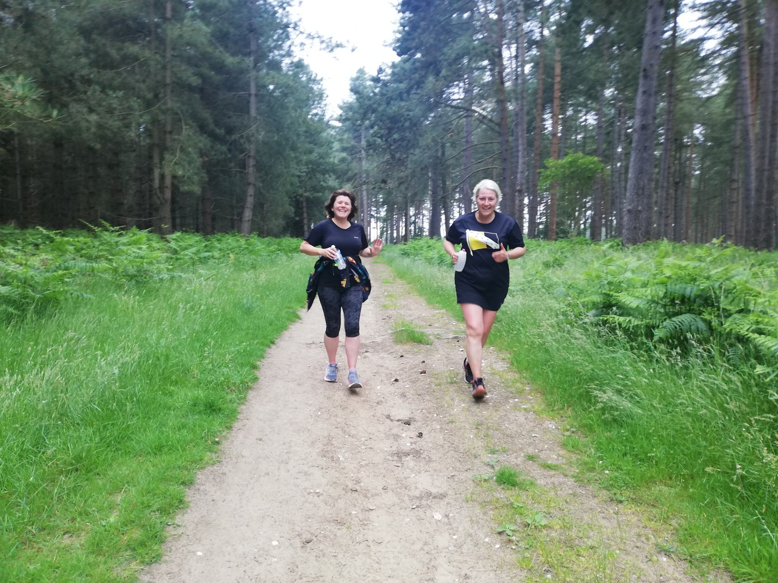 Two women are jogging on a gravel path through a lush, green forest. They are wearing athletic clothing and smiling as they run. Ferns and tall trees surround the path, creating a serene nature scene. The sky is overcast, casting a soft light on the runners.