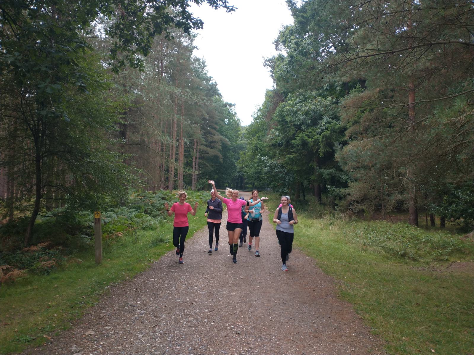 A group of six people jogs together down a wooded trail. They are all dressed in athletic clothing, and the person in the center raises their arms triumphantly. The surrounding trees and greenery suggest a serene forest setting.