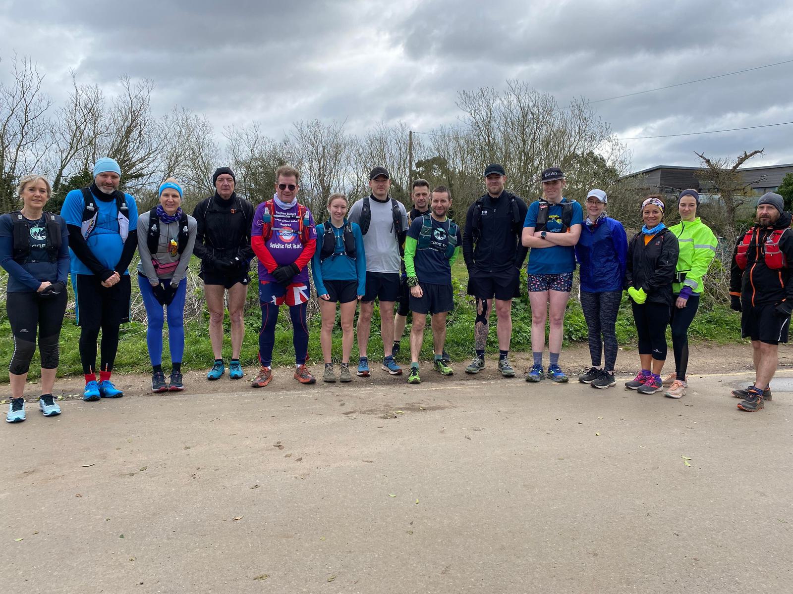 A group of people dressed in athletic gear stand together outdoors on a paved path. The sky is overcast, and there are trees in the background. They appear ready for an outdoor activity, possibly running or walking.