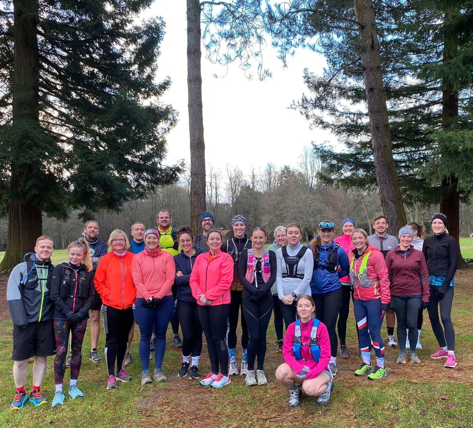 A group of people in athletic wear stand together outdoors, surrounded by tall pine trees. They appear to be in a park or forest setting, posing for a group photo with smiling faces.