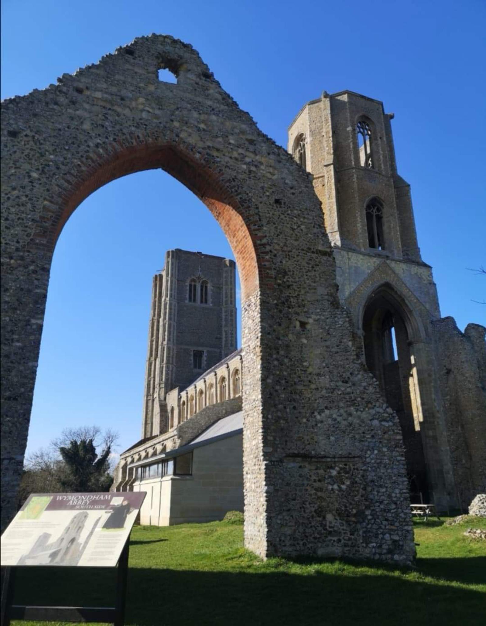 The image shows a historical church ruin featuring a large stone arch and a tall, square bell tower behind it. The structure is made of ancient stone, and an informational sign is visible in the foreground. It's a sunny day with a clear blue sky.