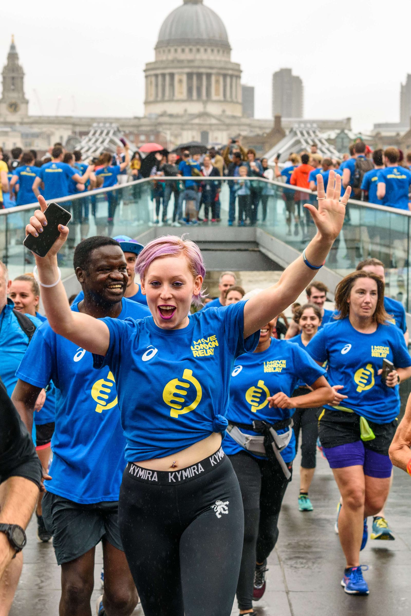 A group of runners in bright blue shirts crosses a bridge during a race. A woman with short pink hair and a phone in her hand leads, raising her arms in excitement. People cheer in the background, and a large domed building is visible in the distant background.