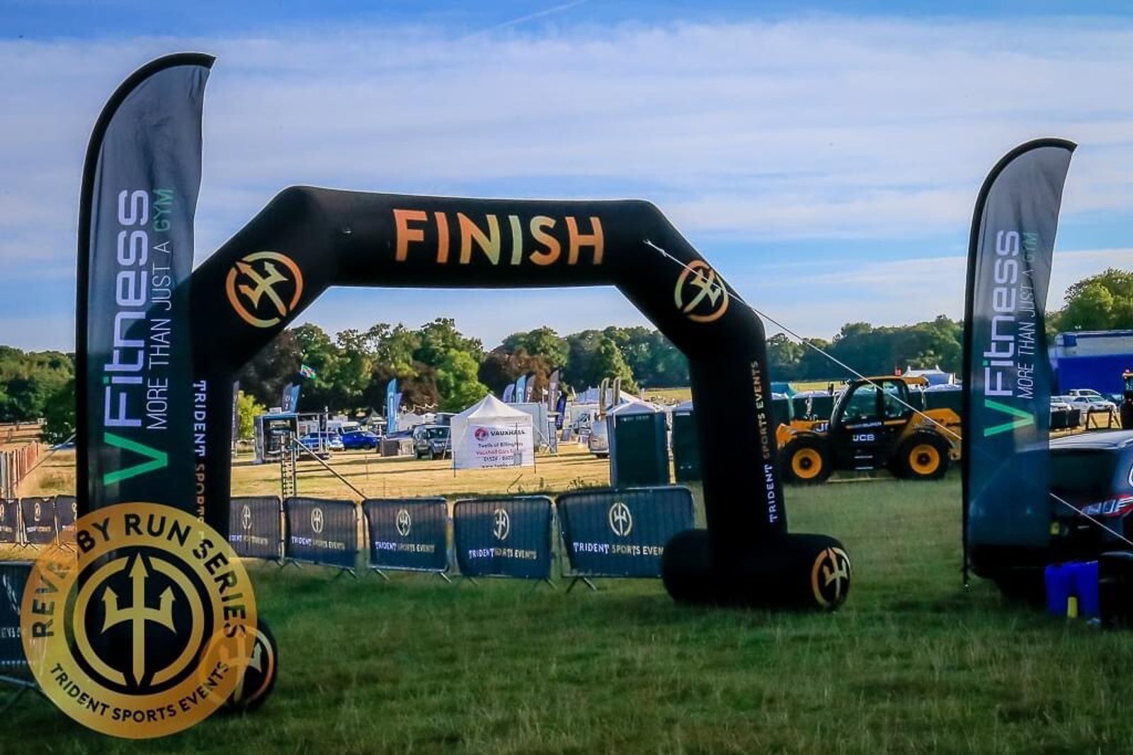 An outdoor finish line at a race event, marked by a black inflatable arch with "Finish" written in orange at the top. There are two black banners on either side displaying event sponsors. In the background are trees, tents, and equipment.
