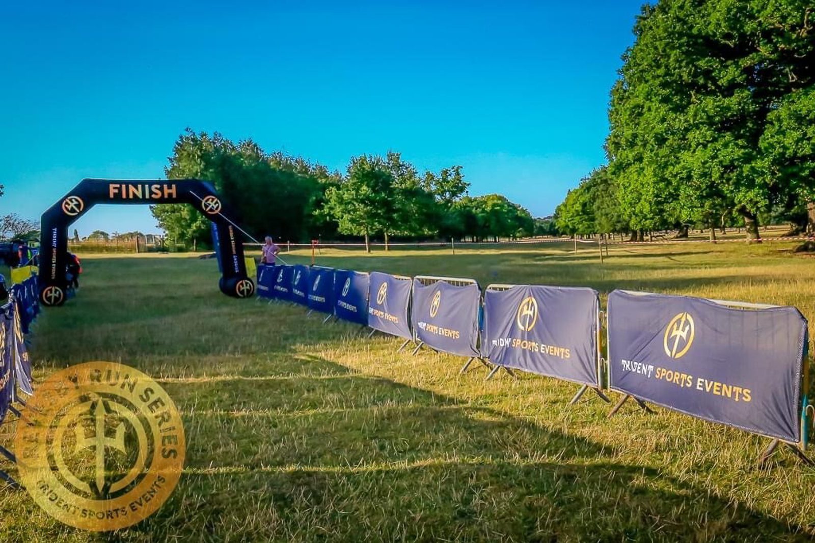 A finish line for a race event set up in a grassy park, with a black and gold inflatable arch marked "FINISH." The pathway is lined with branded barriers featuring the words "TRIDENT SPORTS EVENTS." Trees and a clear blue sky are in the background.