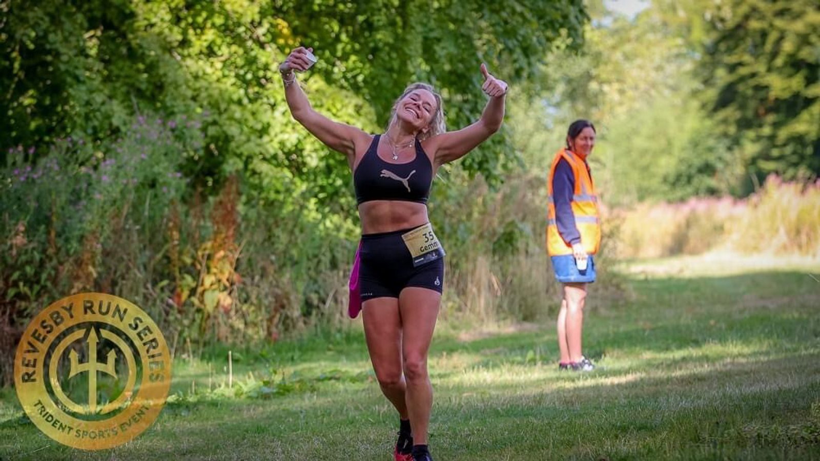 A runner in athletic gear, smiling and giving a thumbs-up, celebrates near the finish line of a race. She wears a yellow race bib and is surrounded by green trees. In the background, a volunteer in an orange vest stands watching. The "Revesby Run Series" logo is in the bottom left corner.